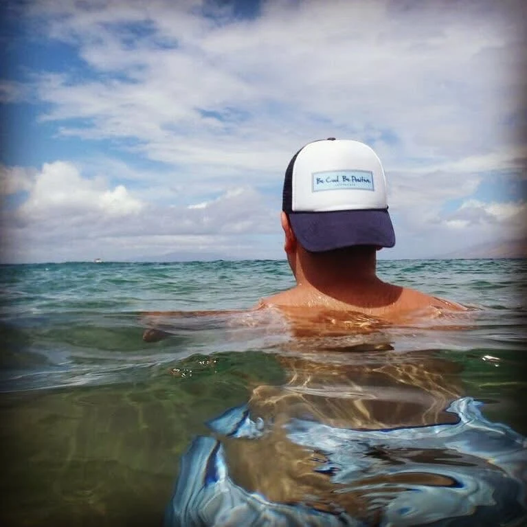The brand's hat, white with a navy brim, worn backwards with the patch to the camera, shoulder deep in clear turquoise ocean under big clouds