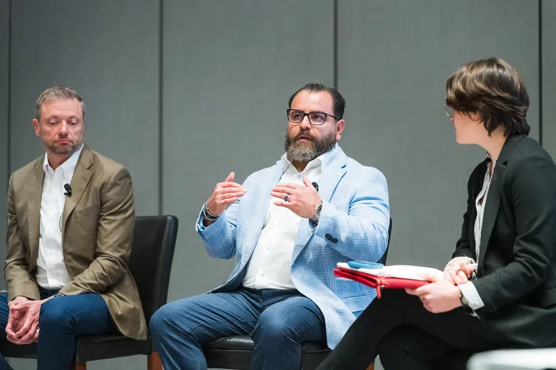Steven F. Gonzalez on a three-person ELEVATE panel, grey room, jeans and a plaid blazer